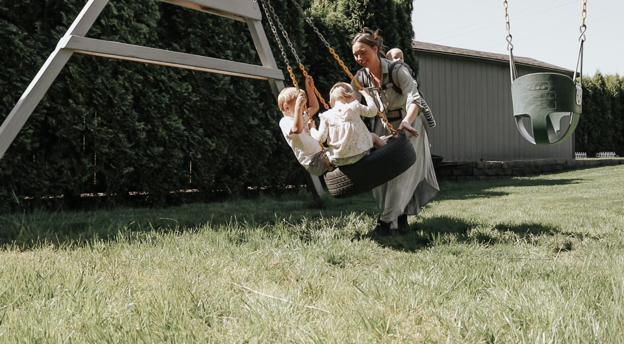 Christian homemaker pushes two children on an outdoor tire swing while carrying a third child in a baby carrier on her back